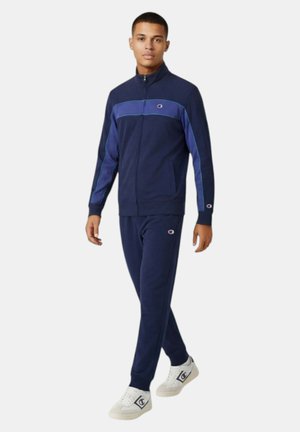Giovane uomo che cammina, indossa una tuta con zip blu navy e viola con scarpe da ginnastica bianche, sfondo chiaro e uniforme.