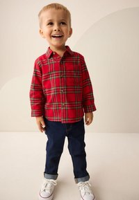 Camicia di flanella a quadri rossi con maniche lunghe e tasca frontale, abbinata a jeans blu scuro risvoltati e scarpe bianche. Tessuto liscio e casual.