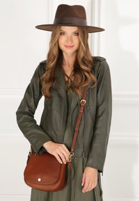 Olivgrüne Leder-Bikerjacke, braune Umhängetasche mit goldenen Hardware-Details und brauner Filzkappe. Glatte Texturen und klare Formen.