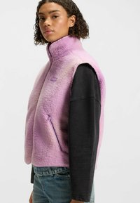 Vaaleanpunainen fleece-liivi, jossa on korkea kaulus, etutasku vetoketjulla ja brodeerattu logo. Musta pitkähiha-paita alla. Rakenteellinen pinta.