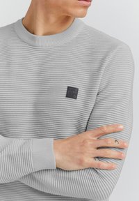 Maglione a coste grigio chiaro con scollatura rotonda; presenta una piccola etichetta nera sul petto e polsini aderenti. Tessuto testurizzato.