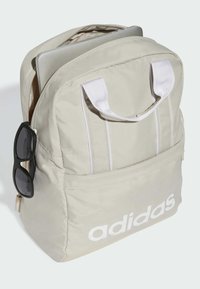 Mochila beige con acentos blancos y gran logo de adidas, que cuenta con un bolsillo frontal y un soporte lateral para gafas de sol, diseñada para portátiles.