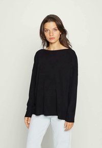 Γυναίκα με κυματιστά καστανά μαλλιά που φορά χρυσά σκουλαρίκια κρίκους, ένα χαλαρό μαύρο μπλουζάκι �με μακριά μανίκια και ανοιχτό μπλε τζιν, μπροστά σε απλό φόντο.