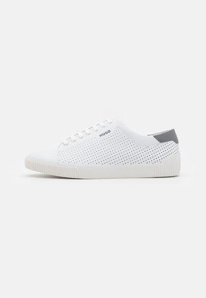 Hvid lavtop-sneaker med perforerede sider, grå hælflap, hvide snørebånd og "HUGO" logo nær snørebåndene på en hvid baggrund.