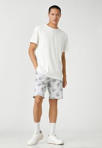 Camiseta blanca de cuello redondo y pantalones cortos grises con patrones de sol. Las zapatillas y los calcetines blancos completan el atuendo. Diseño casual y ajuste relajado.
