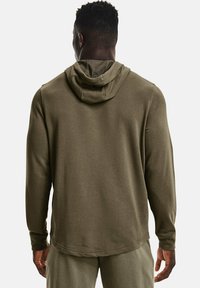 Homme portant un hoodie vert olive et un pantalon assorti, vu de dos contre un fond clair et uni.