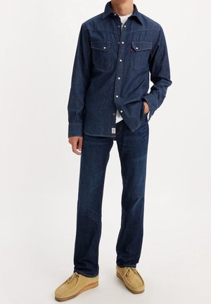 Mężczyzna ubrany w ciemnoniebieską koszulę jeansową z zatrzaskami, pasujące dżinsy, białą podkoszulkę i beżowe zamszowe buty na sznurowadła na białym tle.