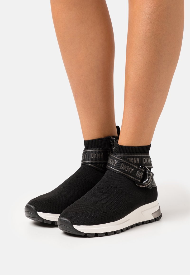 Pieds portant des baskets style chaussettes noires avec semelles blanches et brides noires marquées DKNY autour des chevilles sur fond blanc.