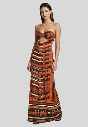 Donna che indossa un maxi abito senza spalline arancione e marrone con motivo geometrico, con spacco frontale e dettaglio ad anello, in piedi davanti a uno sfondo semplice.