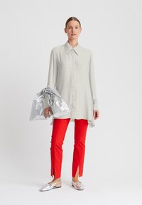 Hellgraues Langarmshirt, rote Slim-Fit-Hose mit seitlichen Schlitzen und silberne Slipper. Das Model hält eine zerknitterte silberne Tasche.