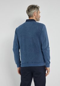Niebieski sweatshirt o teksturowanym wyglądzie z okrągłym dekoltem, ściągaczami na rękawach i u dołu. Wykonany z gładkiego materiału o subtelnym wzorze, noszony z ciemnymi spodniami w drobne paski.