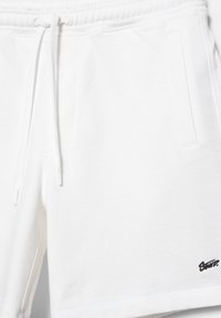 Shorts en coton blanc avec une taille élastique, un cordon de serrage, des poches latérales et un petit logo noir brodé près de l'ourlet.