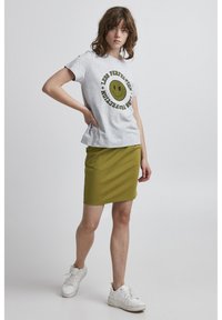 T-shirt en coton gris avec un graphique de visage souriant jaune et du texte, associé à une jupe ajustée vert olive. Des baskets blanches complètent la tenue.