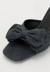 Sandalo con tacco alto in denim, caratterizzato da un grande fiocco sulla parte anteriore, punta quadrata e lati traforati; principalmente di colore grigio scuro con una sottile texture.