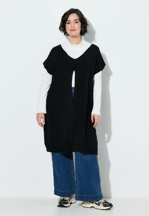 Kvinde med kort mørkt hår iført en lang sort ærmeløs vest over en hvid langærmet top, brede blå jeans og sorte og hvide sneakers.