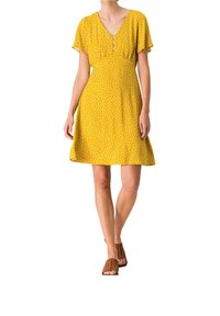 Femme portant une robe jaune à manches courtes avec des pois blancs et des chaussures ouvertes marron, debout sur un fond blanc.