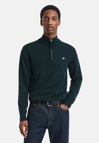 Dunkelgrüner Reißverschluss-Pullover mit hohem Kragen, gerippten Bündchen und kleinem Logo auf der linken Brust, kombiniert mit dunkelblauen Jeans.
