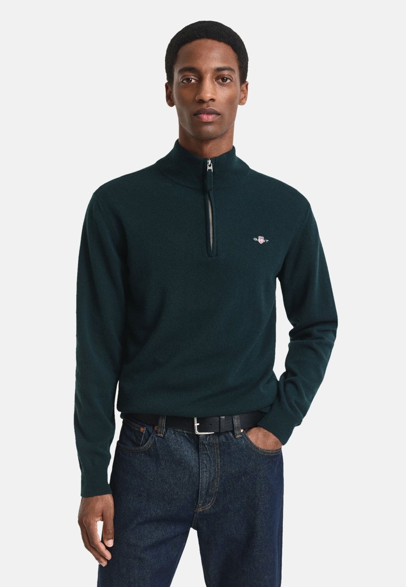 Dunkelgrüner Reißverschluss-Pullover mit hohem Kragen, gerippten Bündchen und kleinem Logo auf der linken Brust, kombiniert mit dunkelblauen Jeans.