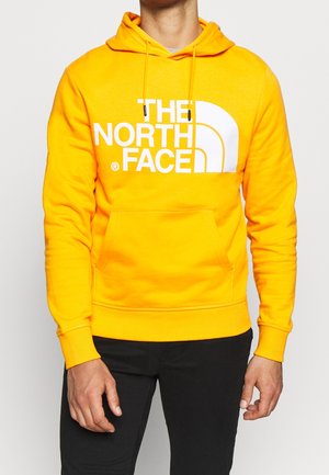 Mand iført en lys gul hættetrøje med frontlomme og stort hvidt "THE NORTH FACE"-logo, kombineret med sorte bukser.