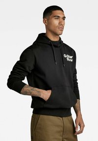 Schwarzer Kapuzenpullover mit Kordelzug, vordertasche im Känguru-Stil, gerippte Bündchen und Saum, mit einem weißen "G-Star Raw"-Logo auf der Brust.