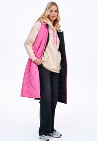 Pikowany, długi kamizelka w jasnoróżowym i czarnym kolorze. Model nosi beżową bluzę z kapturem, ciemne dżinsy i trampki, z akcentowanym różowym wykończeniem.