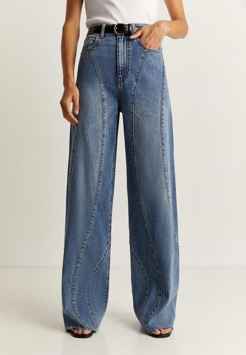 Jean en denim bleu à jambes larges avec détail de couture incurvée, taille haute et poches avant, associé à une ceinture noire et des sandales.