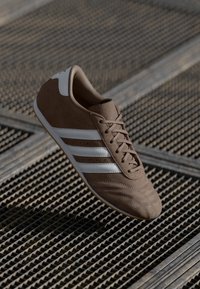 Brown textile sneaker with white stripes, rounded toe, and textured surface. Features laces and a gum sole. Positioned above a metal grid.