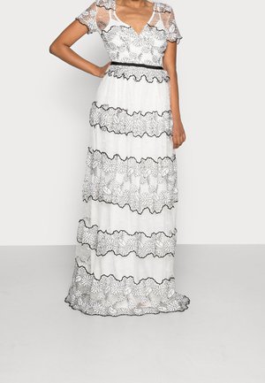 Kvinde iført en hvid maxi kjole med sorte blomstrede blonde mønstre, gennemsigtige korte ærmer og en figursyet talje med sort kant.