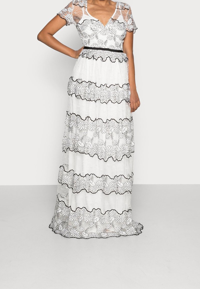 Femme portant une robe maxi blanche avec des motifs floraux en dentelle noire, des manches courtes transparentes et une taille cintrée avec bordure noire.