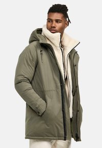 Parka acolchada de color verde olivo con forro de lana crema desmontable, capucha ajustable, cierre frontal con cremallera y bolsillos laterales. Tela texturizada visible.