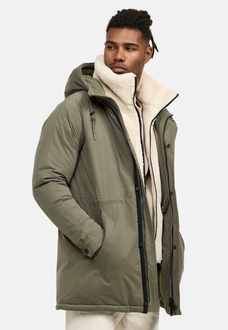 Parka acolchada de color verde olivo con forro de lana crema desmontable, capucha ajustable, cierre frontal con cremallera y bolsillos laterales. Tela texturizada visible.