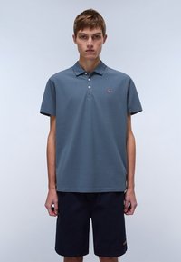 Grå kortermet polo-shirt med krage, tre-knappers knappestel og liten logo detalj. Båret med mørke shorts, frontutsikt.