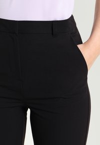 Närbild av en person som bär svarta tajta byxor med bälteshällor, där ena handen är placerad i den högra fickan.