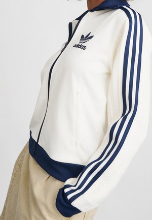 Osoba nosząca białą kurtkę zapinaną na zamek Adidas z granatowymi paskami na rękawach i granatowym obszyciem, w połączeniu z beżowymi spodniami.