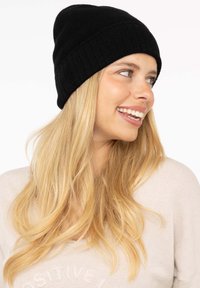 Black knitted beanie hat, fitted design, soft texture, seamless top. Worn on a light-haired individual against a plain background.