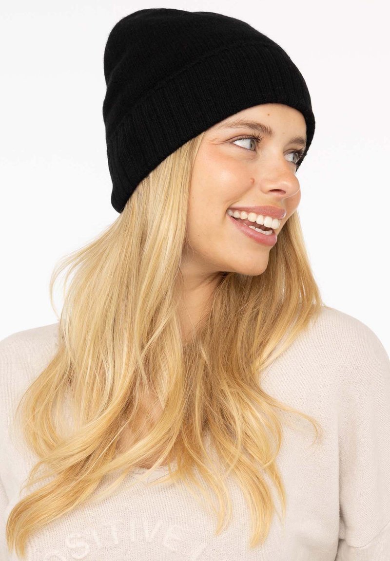 Black knitted beanie hat, fitted design, soft texture, seamless top. Worn on a light-haired individual against a plain background.