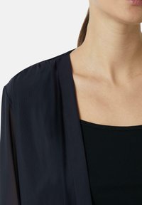 Czarny, lekki kimono o miękkiej, przezroczystej teksturze i otwartym przodzie. Posiada zwisający fason i delikatny połysk, w połączeniu z jednolitym czarnym topem na ramiączkach.