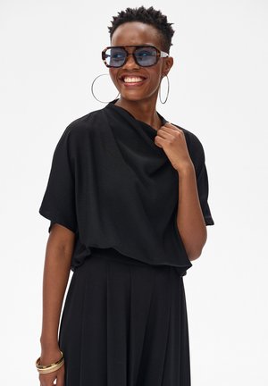 Uśmiechnięta kobieta w dużej czarnej bluzce i plisowanej spódnicy nosi duże kolczyki koła, okulary przeciwsłoneczne w oprawkach przypominających żółwia i złote bransoletki na jednym nadgarstku.