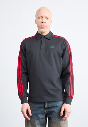 Polo shirt noir à manches longues avec des rayures rouges le long des épaules et des manches, mettant en avant un logo Adidas vert sur la poitrine. Tissu texturé.