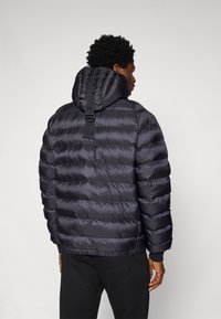 Giacca a vento nera con cappuccio, design trapuntato e una cintura a contrasto al collo. Il materiale ha una texture liscia e lucida.