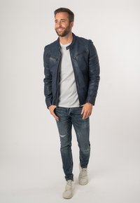 Giacca in pelle blu navy con colletto alto, tasche sul petto con zip, vestibilità slim, abbinata a una t-shirt grigia chiara e jeans blu strappati.