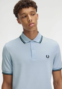 Hellblaues Polohemd mit strukturiertem Finish, schwarz-grün gestreiftem Kragen und kleinem schwarzem Logo auf der linken Brust.