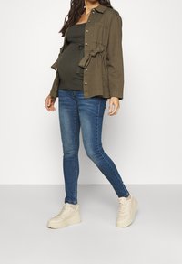 Giacca verde oliva con cintura sopra un top nero, abbinata a jeans skinny blu e sneaker chunky beige. Tessuto morbido, vestibilità casual.