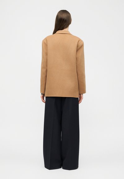 Hosszú barna hajú nő, teve színű kabátot és széles fekete nadrágot visel, háttal áll egy egyszerű, világos háttér előtt.