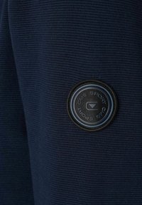 Marineblå tekstureret stof med et rundt sort mærke, hvor logoet "CMS SPORT" er tydeligt vist. Overfladen har fine ribber.