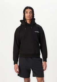 Svart hettegenser med kengurulomme foran. Har en logo på brystet, ribbede mansjetter og nederkant, kombinert med sorte shorts.