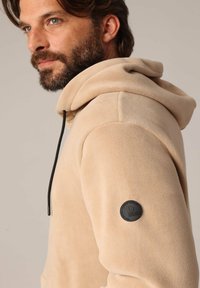 Man met baard in een beige fleece hoodie met zwarte trekkoord en een rond zwart logo op de bovenste mouw, kijkend naar de zijkant.