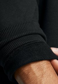 Čierny rukáv mikiny s rebrovanou pleteninou, s viditeľnou mäkkou fleecovou podšívkou. Sú tiež prítomné detaily švov, ktoré zvyšujú odolnosť.