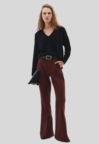 Donna con capelli ricci, che indossa un maglione nero a V, pantaloni larghi marroni, cintura nera, in mano una pochette nera, in piedi su uno sfondo grigio.