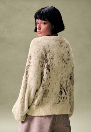 Tânără cu păr negru scurt, purtând un pulover crem pufos cu pete închise, uitându-se peste umăr, pe un fundal verde estompat.